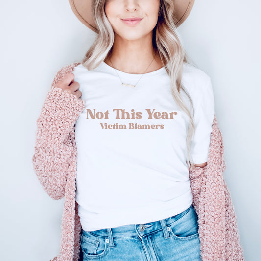 The front of the sweatshirt proudly displays the phrase "Not This Year Victim Blamers" in a bold and eye-catching font. This message serves as a reminder that we reject victim-blaming and support those who have faced injustice.