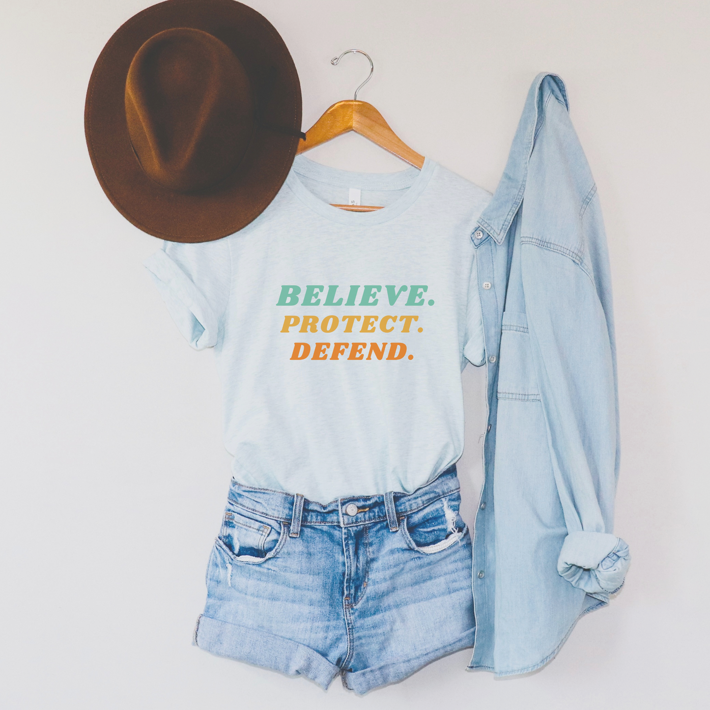 Heather Ice Blue Bella Canvas T-shirt with bold retro text in green, yellow, and orange that reads: Believe. Protect. Defend.