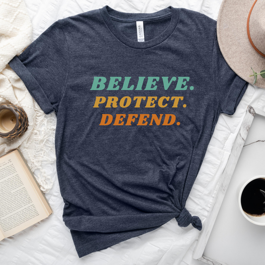 Heather navy blue Bella Canvas T-shirt with bold retro text in green, yellow, and orange that reads: Believe. Protect. Defend.