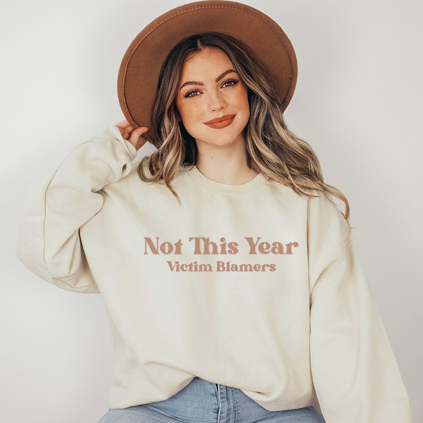 The front of the sweatshirt proudly displays the phrase "Not This Year Victim Blamers" in a bold and eye-catching font. This message serves as a reminder that we reject victim-blaming and support those who have faced injustice.