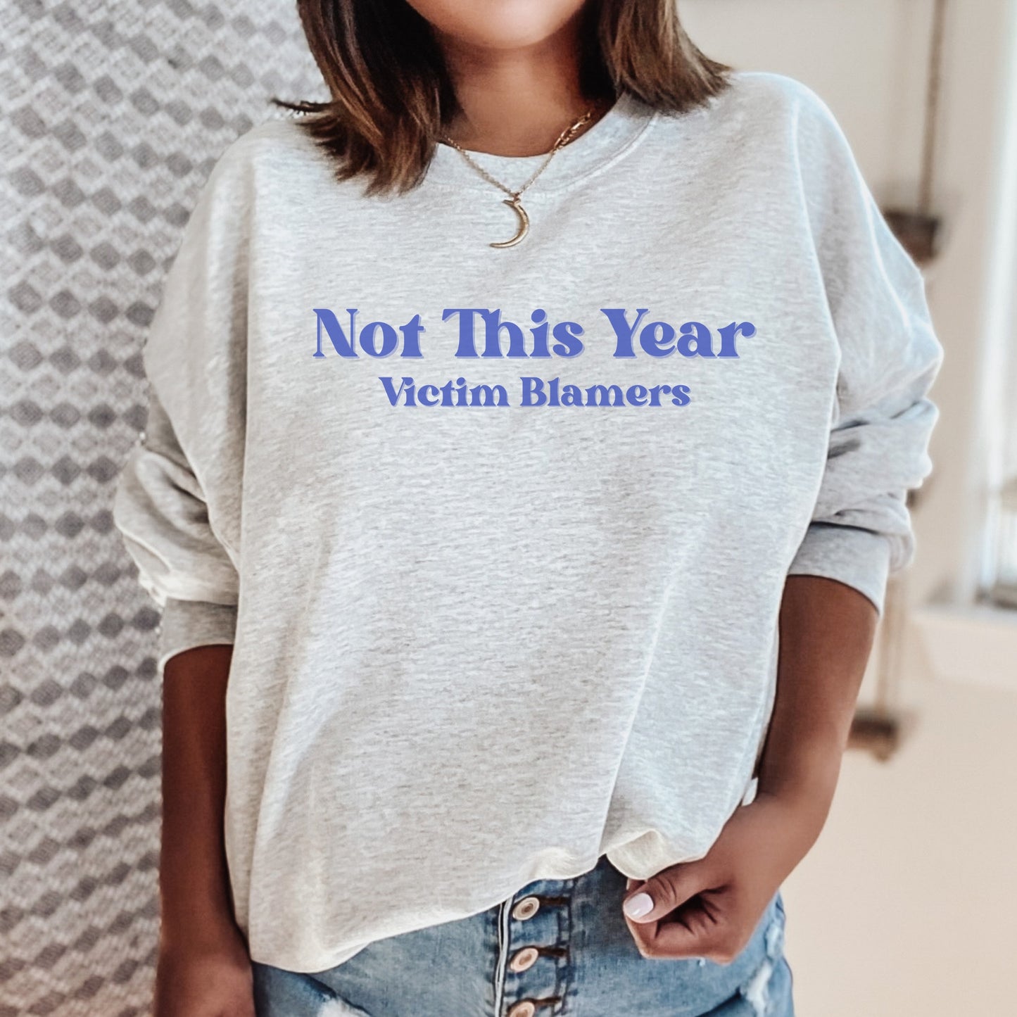 The front of the sweatshirt proudly displays the phrase "Not This Year Victim Blamers" in a bold and eye-catching font. This message serves as a reminder that we reject victim-blaming and support those who have faced injustice.