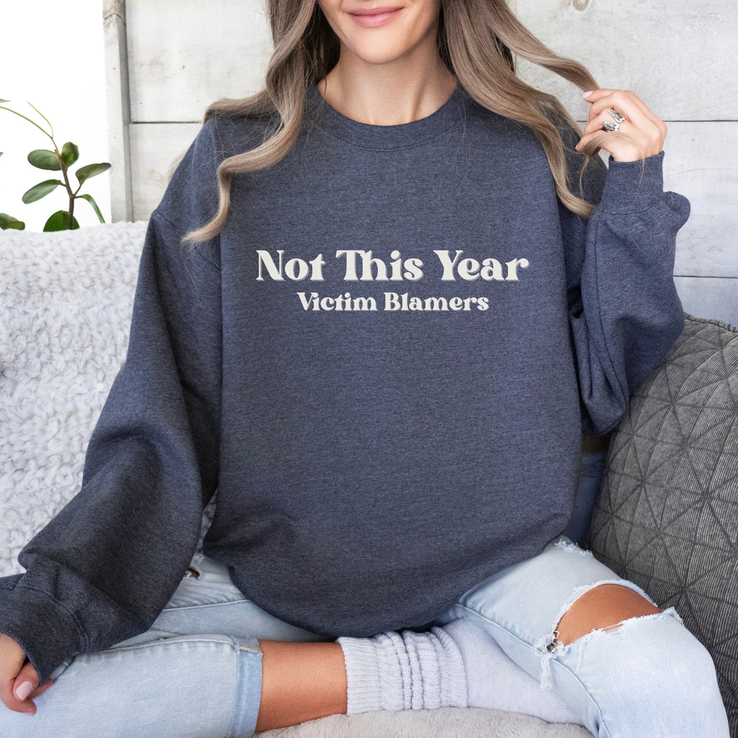 The front of the sweatshirt proudly displays the phrase "Not This Year Victim Blamers" in a bold and eye-catching font. This message serves as a reminder that we reject victim-blaming and support those who have faced injustice.