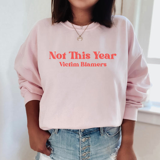 The front of the sweatshirt proudly displays the phrase "Not This Year Victim Blamers" in a bold and eye-catching font. This message serves as a reminder that we reject victim-blaming and support those who have faced injustice.