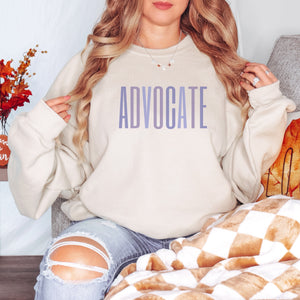 Sand crewneck sweatshirt with a powerful  "ADVOCATE" wordmark emblazoned across the chest in varying shades of purple, symbolizing the strength and unity of domestic violence advocates. The gradient effect from deep violet to lavender adds an eye-catching touch, making this sweater a visually compelling tribute to your commitment to ending domestic abuse.